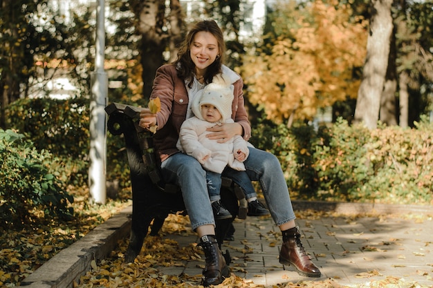 Młoda, słodka matka przytula swoją córkę i pokazuje jej opadły jesienny liść. Mama i dziecko siedzą na ławce w parku