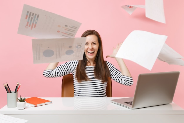 Młoda śmiejąca się kobieta rzuca papierowe dokumenty podczas pracy nad projektem, siedząc w biurze z laptopem przed weekendem na białym tle na różowym tle. Koncepcja kariery biznesowej osiągnięcia. Skopiuj miejsce.