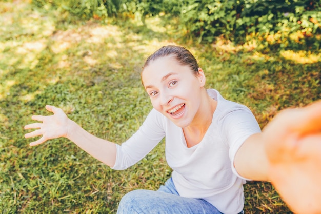 Zdjęcie młoda śmieszna dziewczyna bierze selfie od ręk z telefonu obsiadaniem na zielonej trawy parku lub ogródu tle. portret młoda atrakcyjna kobieta robi selfie fotografii na smartphone w letnim dniu.