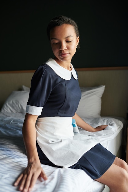 Młoda spokojna pokojówka w mundurze siedzi na czystym łóżku w pokoju hotelowym