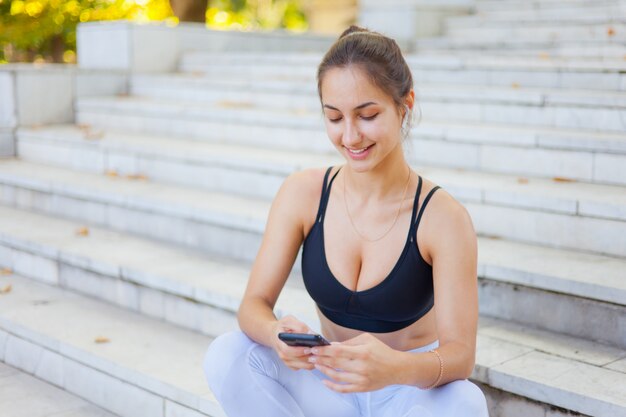 Młoda sport kobieta w sportowym używa smartphone i słucha muzyka w hełmofonach podczas gdy siedzący na schodkach w jaskrawym słonecznym dniu