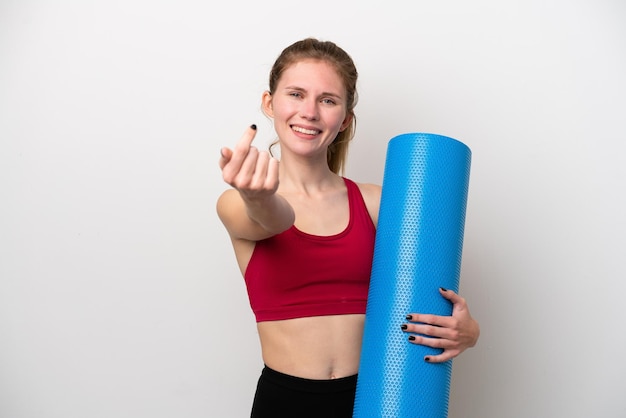 Młoda sportowa Angielka idzie na zajęcia jogi, trzymając matę na białym tle, wykonując gest nadchodzący