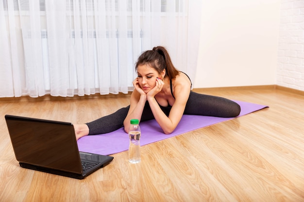 Młoda sportowa kobieta ćwiczy jogę na fioletowej macie i patrzy na swojego laptopa