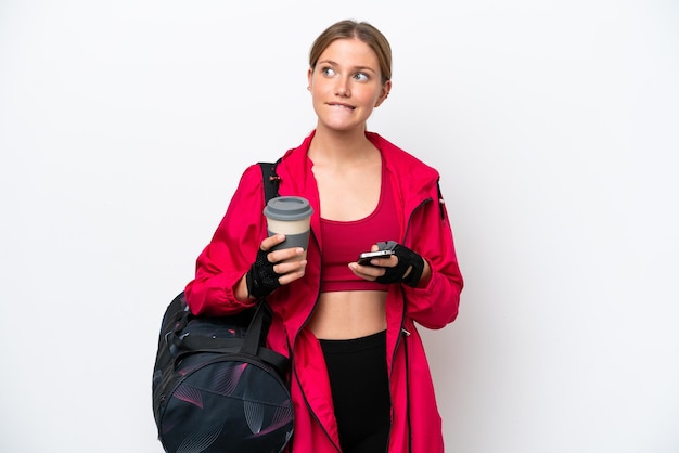 Młoda sportowa kobieta kaukaska na białym tle trzymająca kawę na wynos i telefon komórkowy, myśląc o czymś