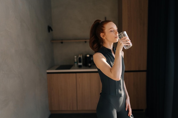 Młoda, sportowa kobieta w odzieży sportowej stoi w domowej kuchni i pije świeżą wodę po zajęciach.