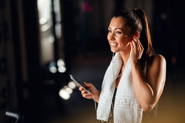 Młoda sportsmenka korzystająca z telefonu komórkowego i słuchająca muzyki przez słuchawki podczas treningu sportowego na siłowni