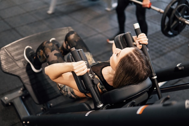 Młoda sprawna kobieta robi ćwiczenia nóg przy użyciu maszyny treningowej w siłowni. Widok z tyłu