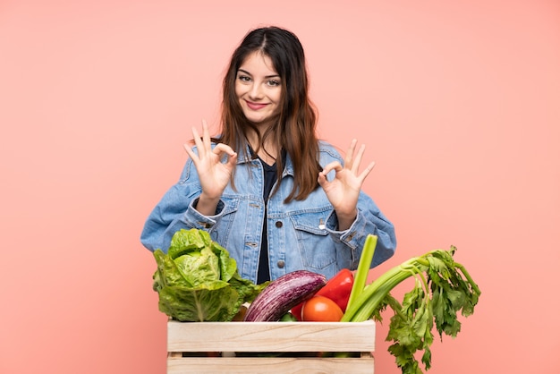 Młoda średniorolna kobieta trzyma kosz pełno świezi warzywa pokazuje ok znaka z palcami
