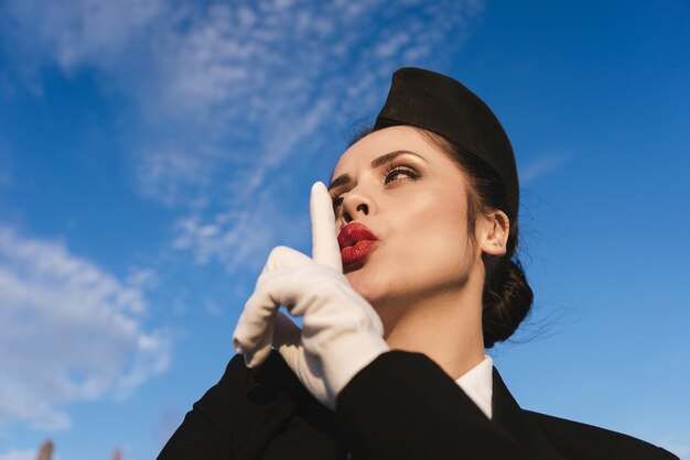 Młoda stewardessa w mundurze, przyłożyła palec do ust, pod błękitnym niebem