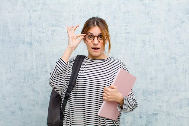 Młoda studencka kobieta czująca się zszokowana, zdziwiona i zaskoczona, trzymająca okulary o zdumionym, niedowierzającym spojrzeniu na ścianie grunge