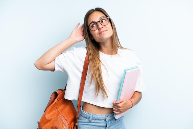 Młoda studencka kobieta odizolowana na niebieskim tle słucha czegoś, kładąc rękę na uchu