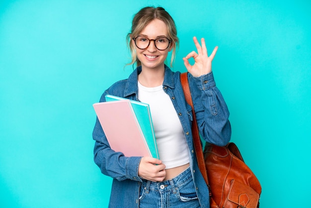 Zdjęcie młoda studencka kobieta odizolowywająca na błękitnym tle pokazuje ok znaka z palcami