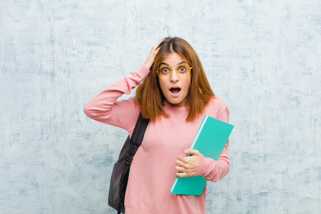 Młoda studencka kobieta wyglądająca na podekscytowaną i zaskoczoną, z otwartymi ustami obiema rękami na głowie, czująca się jak szczęśliwy zwycięzca