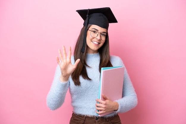 Młoda studentka brazylijska kobieta w kapeluszu z podziałką na różowym tle, licząc pięć palcami