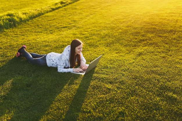 Młoda studentka inteligentna kobieta w ubranie. Kobieta leży na ziemi trawy, pracując na komputerze przenośnym w parku miejskim na zielonym trawniku słonecznym na zewnątrz. Biuro mobilne. Koncepcja biznesowa niezależnego.