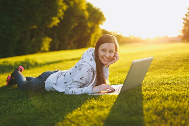 Młoda studentka inteligentna kobieta w ubranie. Kobieta leży na ziemi trawy, pracując na komputerze przenośnym w parku miejskim na zielonym trawniku słonecznym na zewnątrz. Biuro mobilne. Koncepcja biznesowa niezależnego.