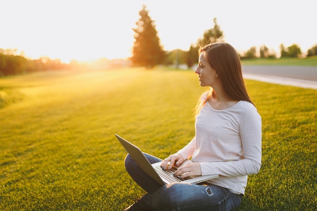 Młoda Studentka Inteligentna Kobieta W Ubranie. Kobieta Siedzi Na Ziemi Trawy, Pracuje Na Komputerze Typu Laptop W Parku Miejskim Na Zielonym Trawniku Słonecznym Na Zewnątrz. Biuro Mobilne. Koncepcja Biznesowa Freelancer