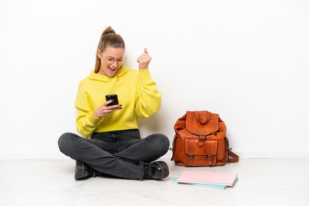 Młoda studentka kaukaska kobieta siedzi na podłodze na białym tle z telefonem w pozycji zwycięstwa