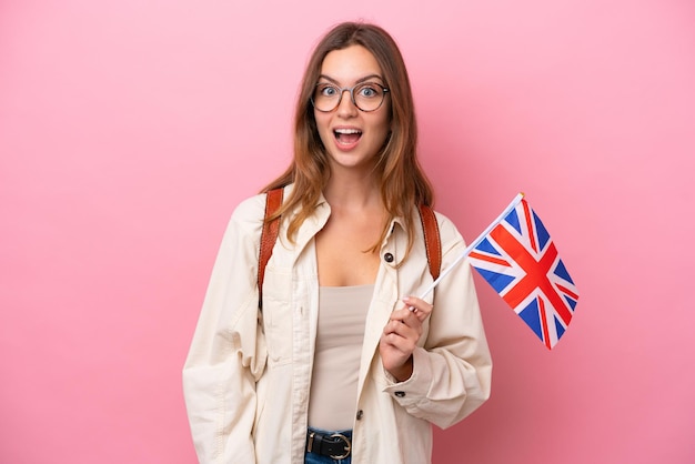 Młoda studentka kaukaska kobieta trzymająca flagę Wielkiej Brytanii na białym tle na różowym tle z niespodzianką wyrazem twarzy