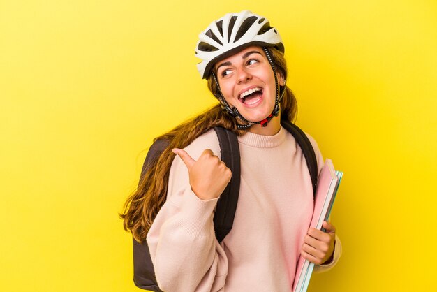 Młoda studentka kaukaski kobieta nosi kask rowerowy na białym tle na żółtym tle wskazuje palcem kciuka, śmiejąc się i beztrosko.