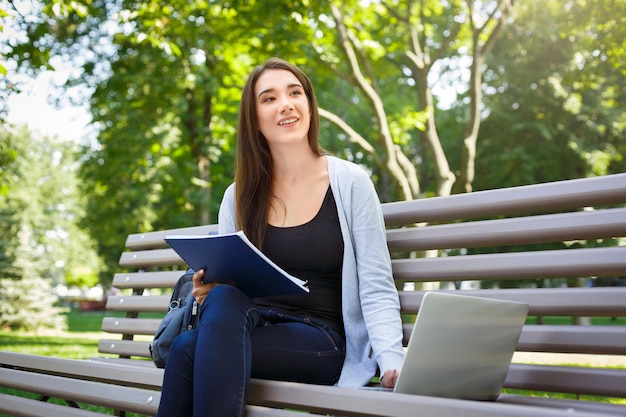 Zdjęcie młoda studentka na ławce. uśmiechnięta brunetka dziewczyna odrabianiu lekcji w parku z laptopem i podręcznikiem. koncepcja stylu życia, technologii i edukacji