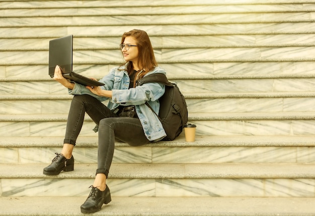 Zdjęcie młoda studentka nowoczesnej kobiety w dżinsowej kurtce i okularach, siedząc na schodach z laptopem. oglądać wideo. nauka na odległość. nowoczesna koncepcja młodzieży.
