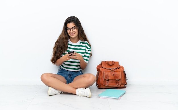 Młoda studentka siedząca na podłodze na białym tle, wysyłająca wiadomość za pomocą telefonu komórkowego