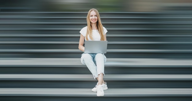 Młoda Studentka Siedzi Z Laptopem Na Schodach W Pobliżu Uniwersytetu, Uśmiechając Się Do Pracy I Patrz Na Kamerę
