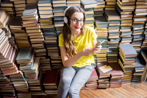 Młoda studentka słuchająca muzyki siedząca na stosie książek w starej bibliotece