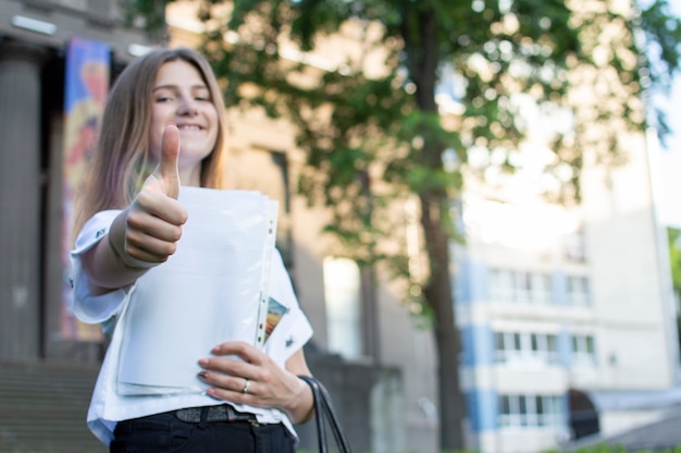 Młoda studentka stojąca w pobliżu uniwersytetu, z uśmiechem w ręku kartką i pokazująca jak na studiach, idzie do szkoły