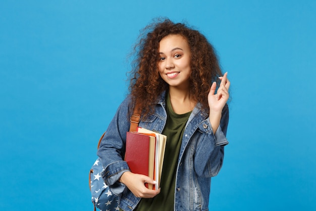 Młoda studentka w dżinsowych ubraniach i plecaku trzyma książki izolowane na niebieskiej ścianie