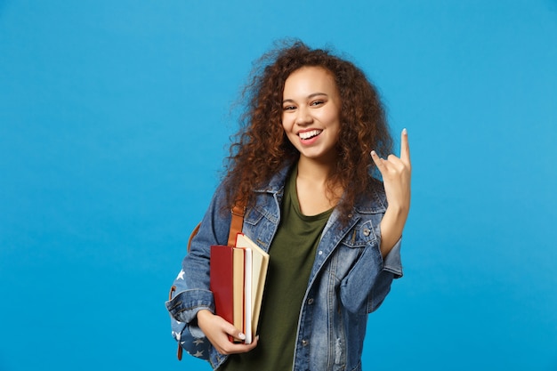 Młoda studentka w dżinsowych ubraniach i plecaku trzyma książki izolowane na niebieskiej ścianie