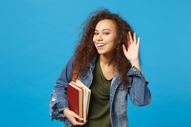 Młoda studentka w dżinsowych ubraniach i plecaku trzyma książki, próbując usłyszeć cię na białym tle na niebieskim tle portret studyjny