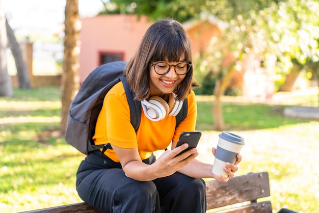 Młoda studentka wygrywa park, używając telefonu komórkowego i trzymając kawę na wynos