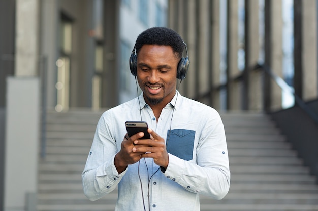 Młoda studentka z Afryki słuchająca muzyki ze smartfona za pomocą dużych słuchawek, uśmiechnięta z wygody korzystania z aplikacji