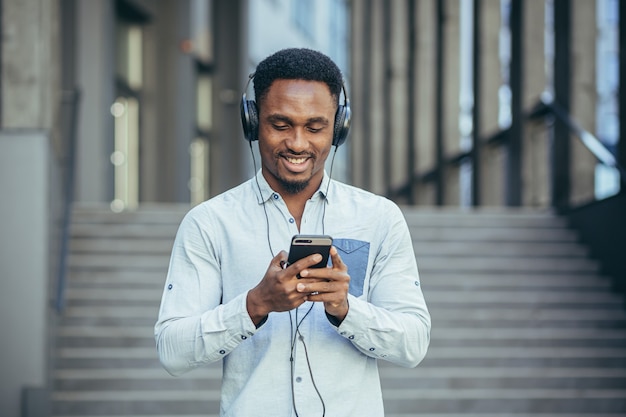Młoda studentka z Afryki słuchająca muzyki ze smartfona za pomocą dużych słuchawek, uśmiechnięta z wygody korzystania z aplikacji