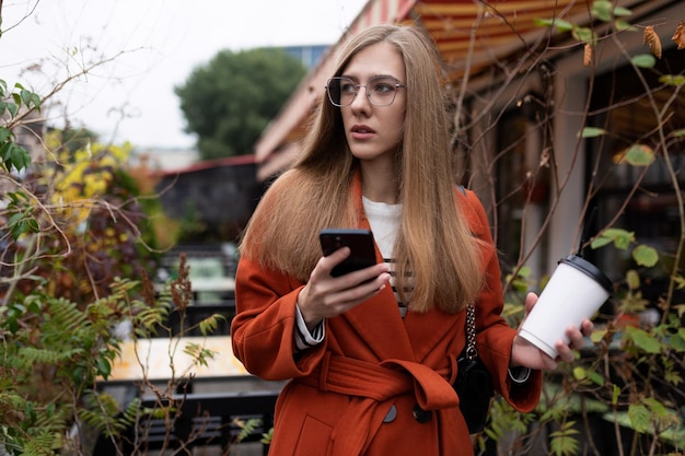 Młoda stylowa bizneswoman z filiżanką kawy w dłoniach i telefonem komórkowym na ulicy w an