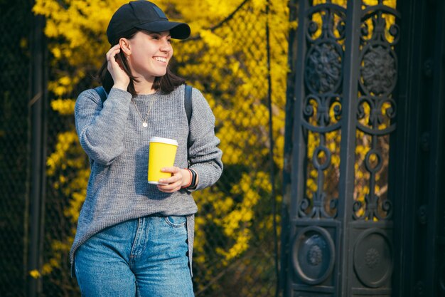 Młoda stylowa kobieta w czapce pijąca kawę na zewnątrz z żółtego papierowego kubka