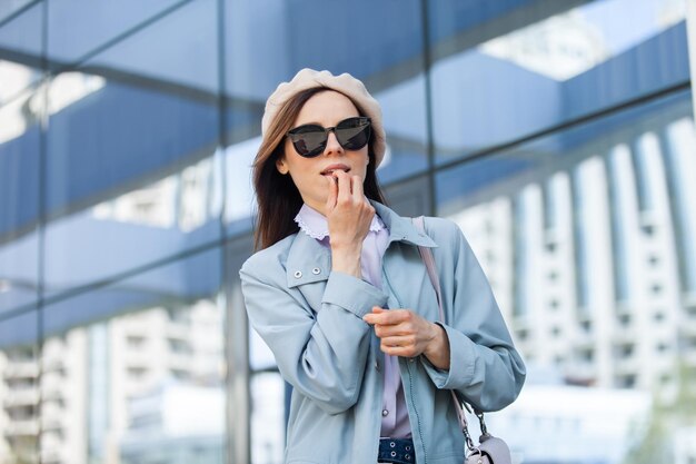 Młoda stylowa kobieta w okularach przeciwsłonecznych i berecie maluje usta na tle okien centrum biznesowego Moda uroda styl życia