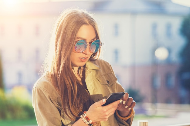 Młoda stylowa ładna kobieta ręce trzymając telefon Zachód słońca w tle Słoneczny dzień, dobra pogoda okulary przeciwsłoneczne fajne akcesoria