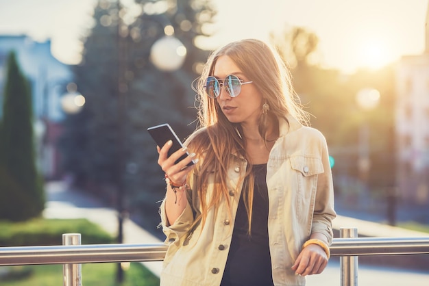 Młoda stylowa ładna kobieta ręce trzymając telefon Zachód słońca w tle Słoneczny dzień, dobra pogoda okulary przeciwsłoneczne fajne akcesoria