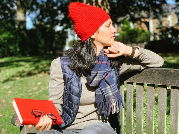 Zdjęcie młoda, szczera kobieta w zwykłych jesiennych ubraniach siedząca na drewnianej ławce w parku i czytająca czerwoną książkę