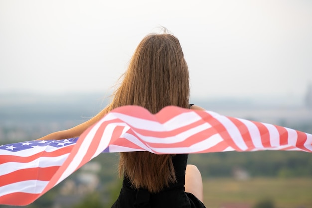 Młoda Szczęśliwa Amerykanka Z Długimi Włosami Trzymająca Macha Na Wietrze Flaga Narodowa Usa Na Jej Sholders Relaks Na świeżym Powietrzu, Ciesząc Się Ciepłym Letnim Dniem