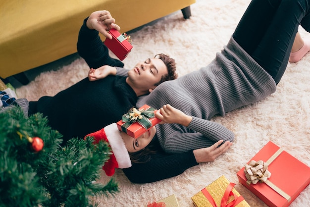 Młoda szczęśliwa azjatycka para ubrana w sweter leżący razem i trzymająca prezenty z wieloma pudełkami na dywanie w salonie podczas świąt Bożego Narodzenia