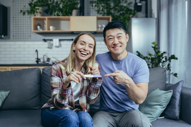 Młoda szczęśliwa azjatycka rodzina i kobieta patrząca w kamerę i radująca się uśmiechając się dostając pozytywny test ciążowy siedząc w domu na kanapie