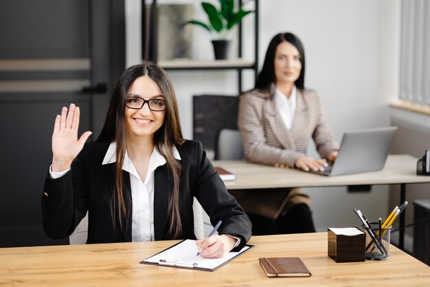 Młoda szczęśliwa bizneswoman używająca notebooka w nowoczesnym biurze z kolegami Dziewczyna w garniturze biznesowym patrząca na kamerę i robiąca gest dłoni na pozdrowienie