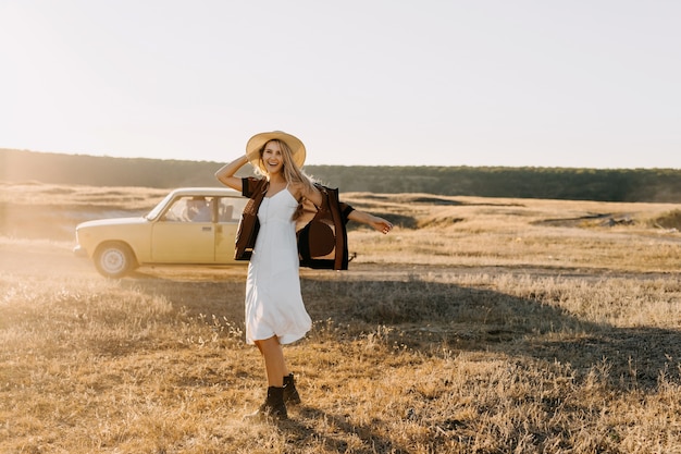 Młoda szczęśliwa blondynka, ubrana w vintage sukienkę i słomkowy kapelusz, chodzenie po polu z suchą trawą