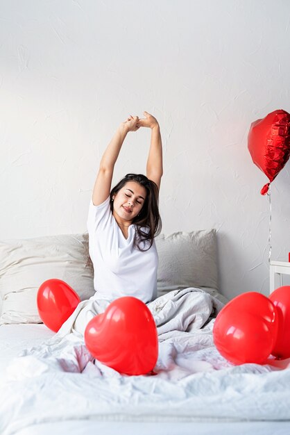 Młoda szczęśliwa brunetka kobieta obudzona w łóżku z czerwonymi balonami w kształcie serca