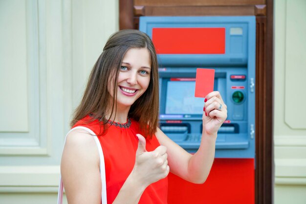 Młoda, szczęśliwa, brunetka wyciąga pieniądze z karty kredytowej w bankomacie.