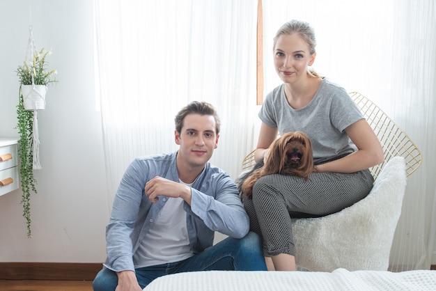 Młoda szczęśliwa caucasian para z uśmiechem siedzi na kanapy uściśnięcia psie na kolanie w sypialni w domu, indoors stylu życia pojęcie.